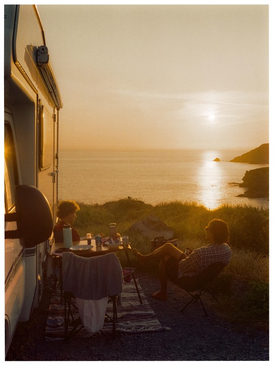 A man and a boy sit at a table next to a camper van, looking out at the ocean at sunset.
