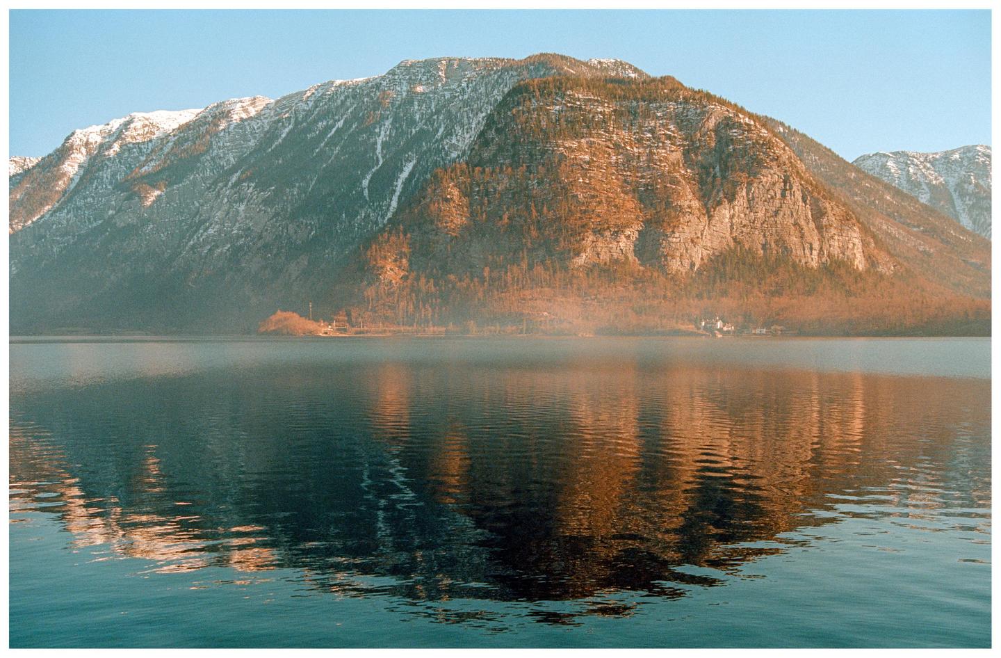 A large mountain with patches of snow and trees rises above a lake with a small village on its shore.