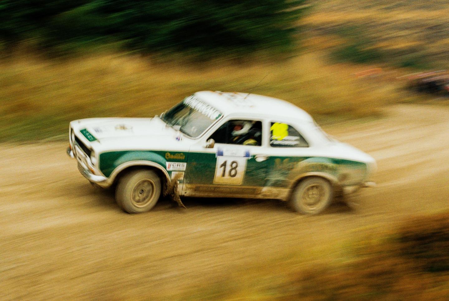 A white and green rally car with the number 18 on its side speeds along a dirt track.
