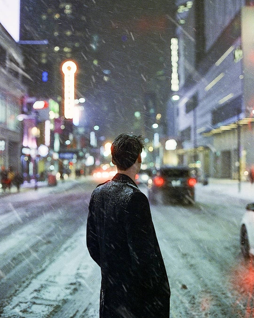 A man stands in the middle of a snowy city street at night, looking away from the camera.