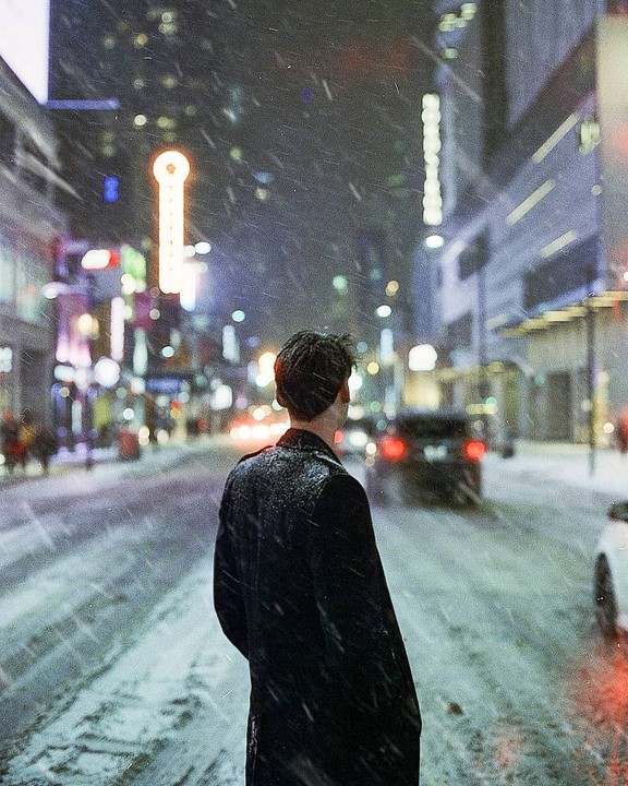 A man stands in the middle of a snowy city street at night, looking away from the camera.