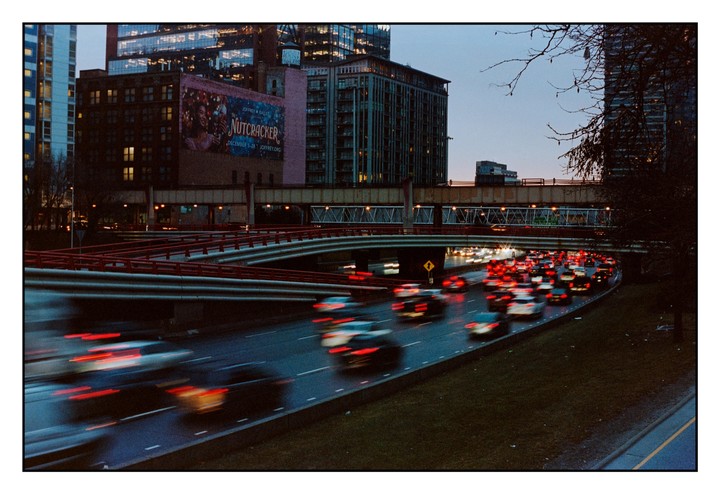 A long exposure shot captures the motion blur of cars on a highway during rush hour, with city buildings and a "Nutcracker" billboard in the background.