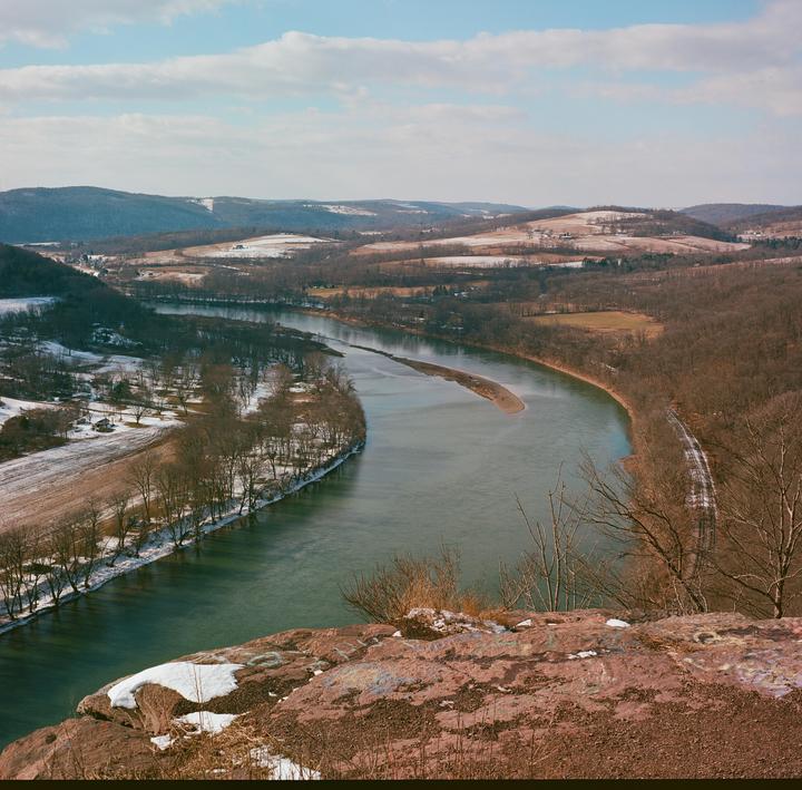 A wide river with a sandbar in the middle flows through a valley with snow-dusted fields and bare trees, viewed from a rocky cliff.