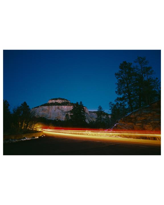 Light trails from vehicles illuminate a road winding through a mountainous landscape at blue hour.