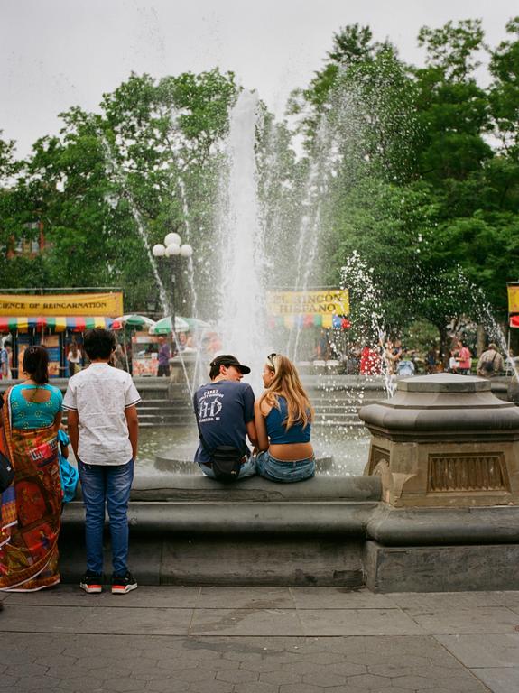 A couple sits on the edge of a fountain while two people stand with their backs to the viewer.