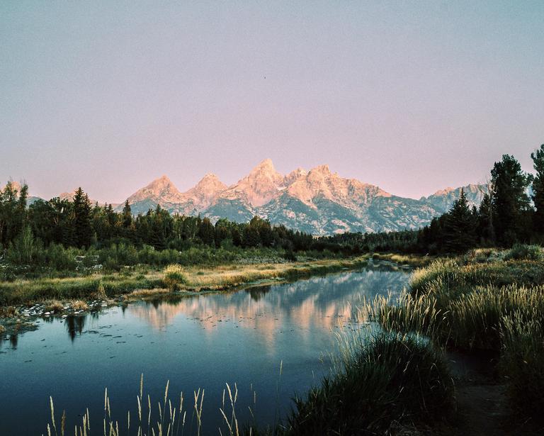 A river reflects mountains and trees under a pink sky.