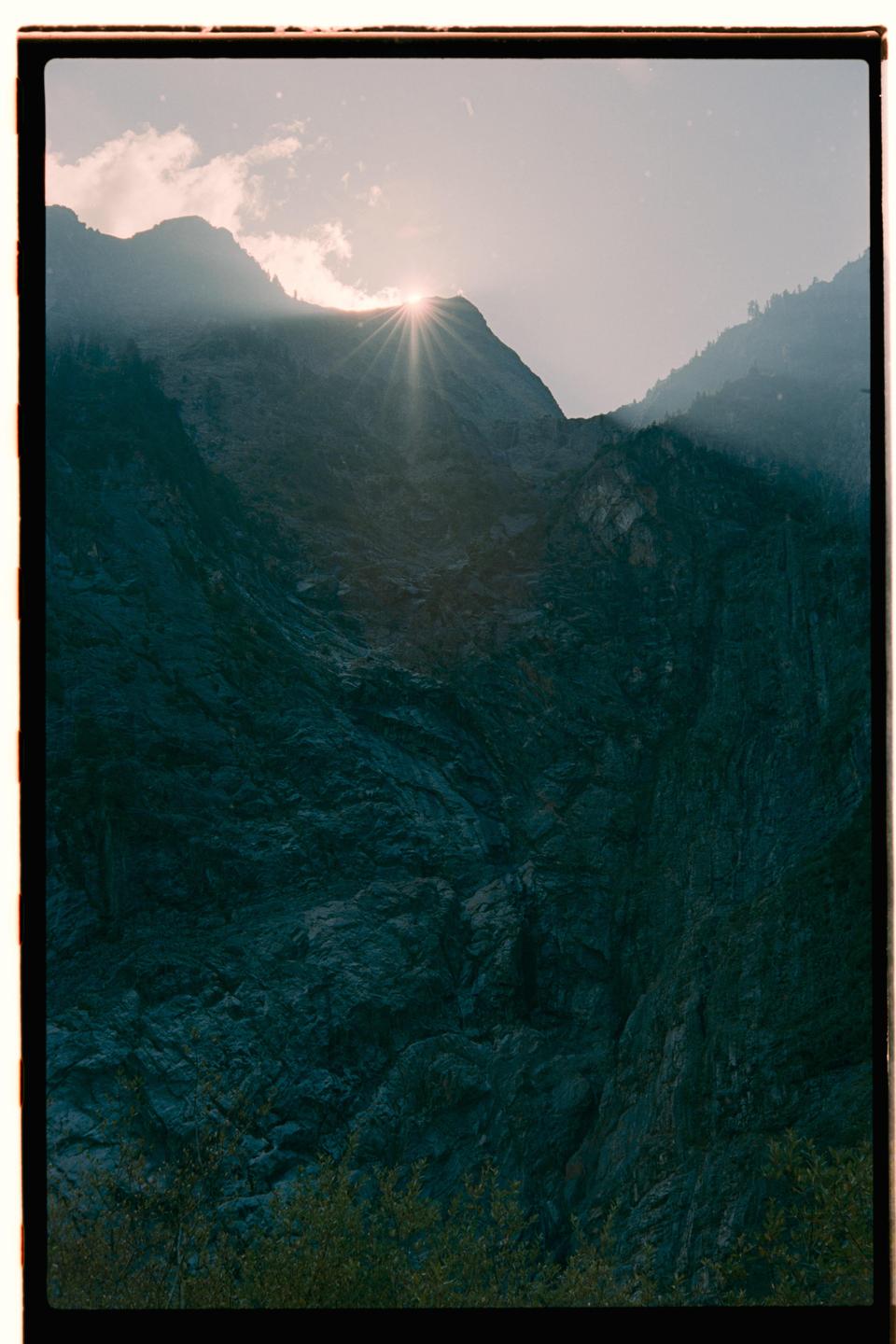 Sunlight bursts over a mountain peak, illuminating the rugged terrain and a hazy sky.
