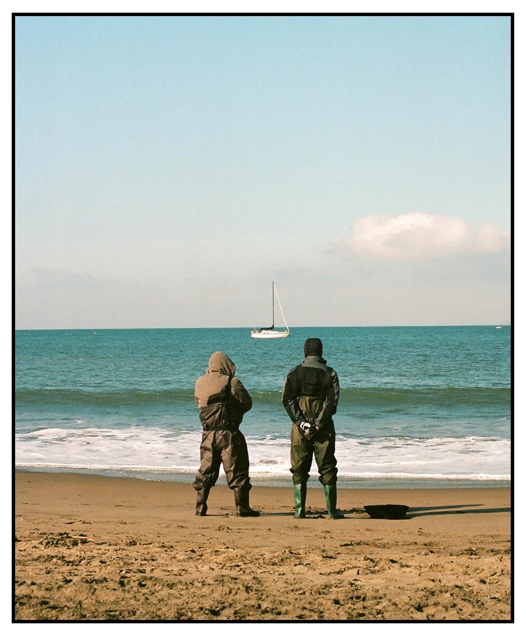 Two men in waders stand on a sandy beach looking out at a sailboat on the ocean.