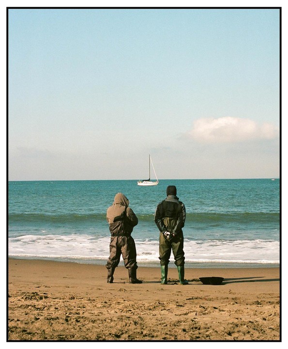 Two men in waders stand on a sandy beach looking out at a sailboat on the ocean.