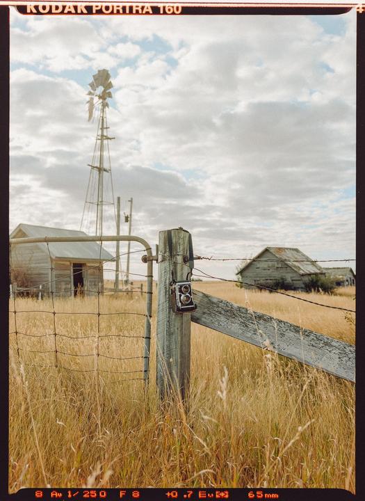 A vintage camera hangs on a wooden fence post in a field with a windmill and old buildings under a cloudy sky.