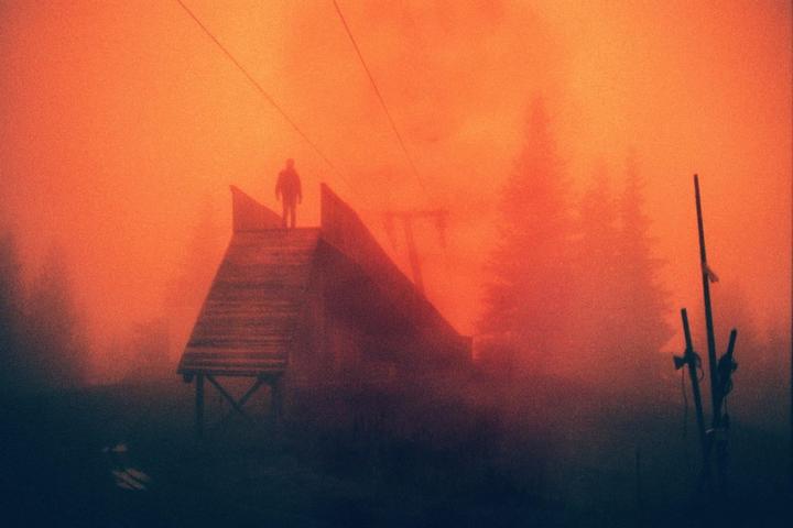 A person stands atop a wooden structure in a hazy, orange-hued landscape with trees and ski equipment visible.