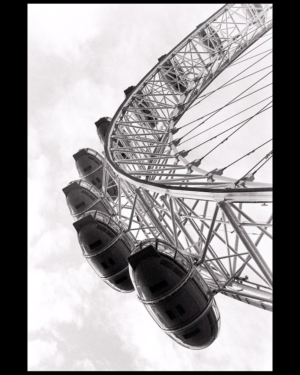 A low-angle black and white shot captures the intricate structure of a ferris wheel against a cloudy sky.