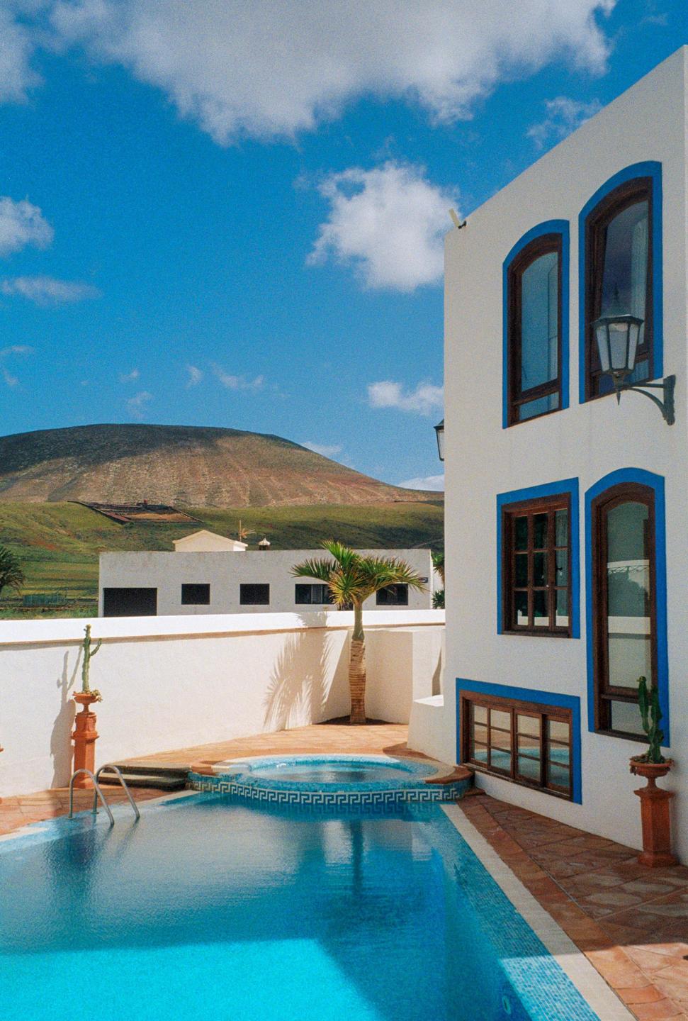 A white building with blue trim and brown windows stands next to a swimming pool and hot tub, with a mountain in the background.