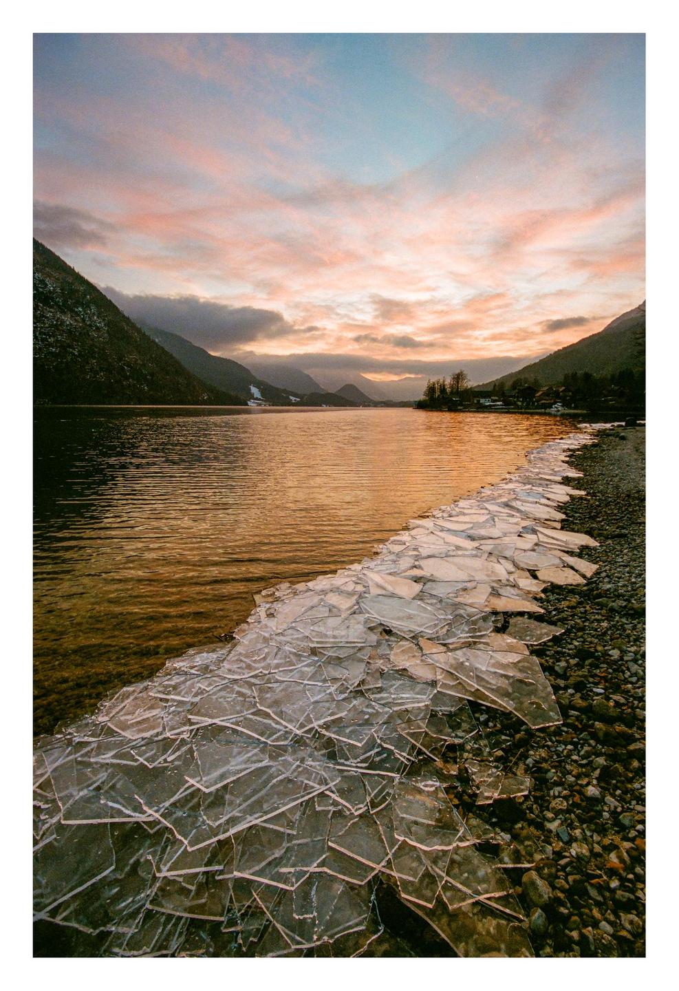 Broken ice sheets are piled on a rocky shore next to a lake with mountains in the background under a colorful sunset sky.