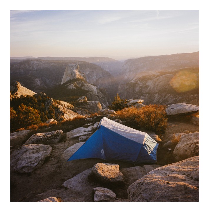 A blue and white tent is pitched on a rocky outcrop overlooking a vast mountain valley with Half Dome visible in the distance.