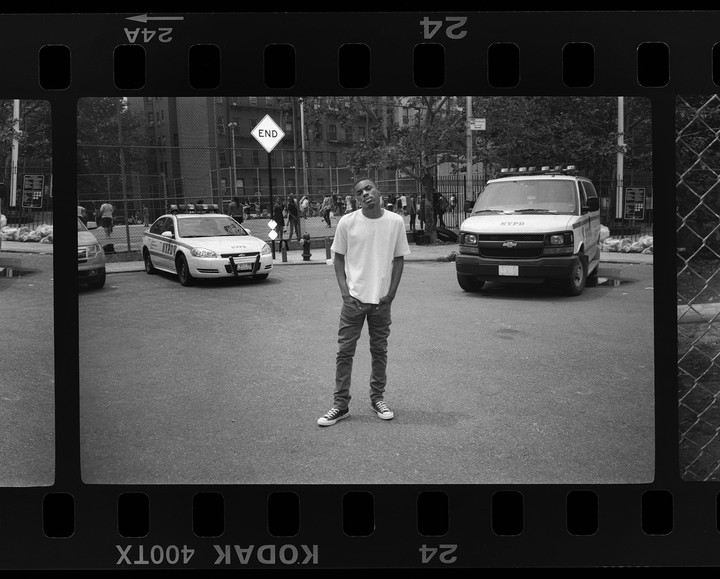 A man stands in the middle of a street with two police vehicles and a chain-link fence in the background.