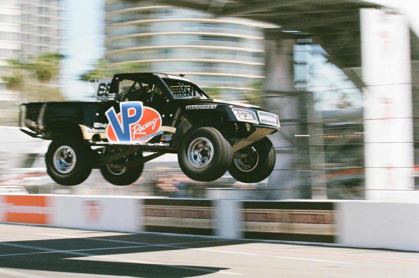A black racing truck with "VP Racing" on its side is captured mid-air above a race track.