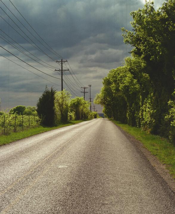 A long, empty road stretches into the distance, flanked by green trees and utility poles under a dramatic, cloudy sky.