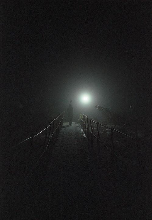 A person walks on a narrow path with railings on both sides, illuminated by a bright light in the distance.