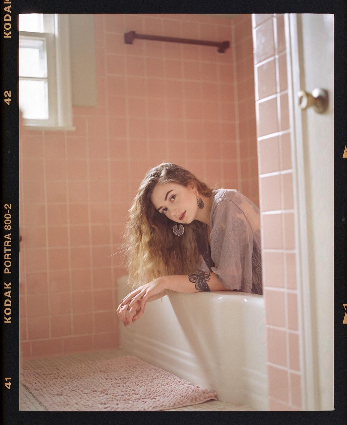 A woman with long, curly hair leans over the edge of a bathtub in a pink-tiled bathroom.