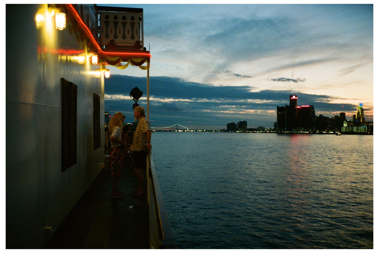A couple stands on the deck of a boat with a city skyline and bridge in the background at dusk.