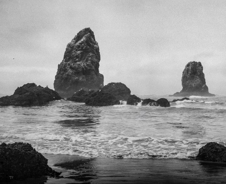 Waves crash on a rocky beach with large sea stacks under a hazy sky.