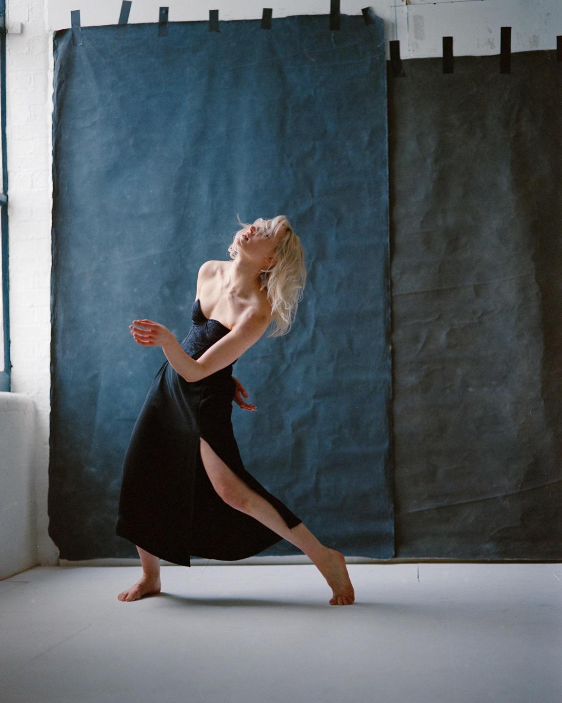 A woman with blonde hair and a black dress is dancing barefoot in a studio with a blue backdrop.