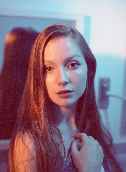 A woman with long hair looks directly at the viewer, illuminated by split lighting in warm and cool tones.