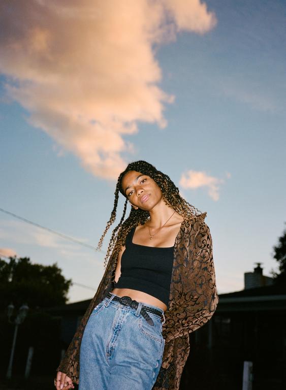 A woman with braided hair and a patterned jacket stands against a blue sky with clouds at sunset.