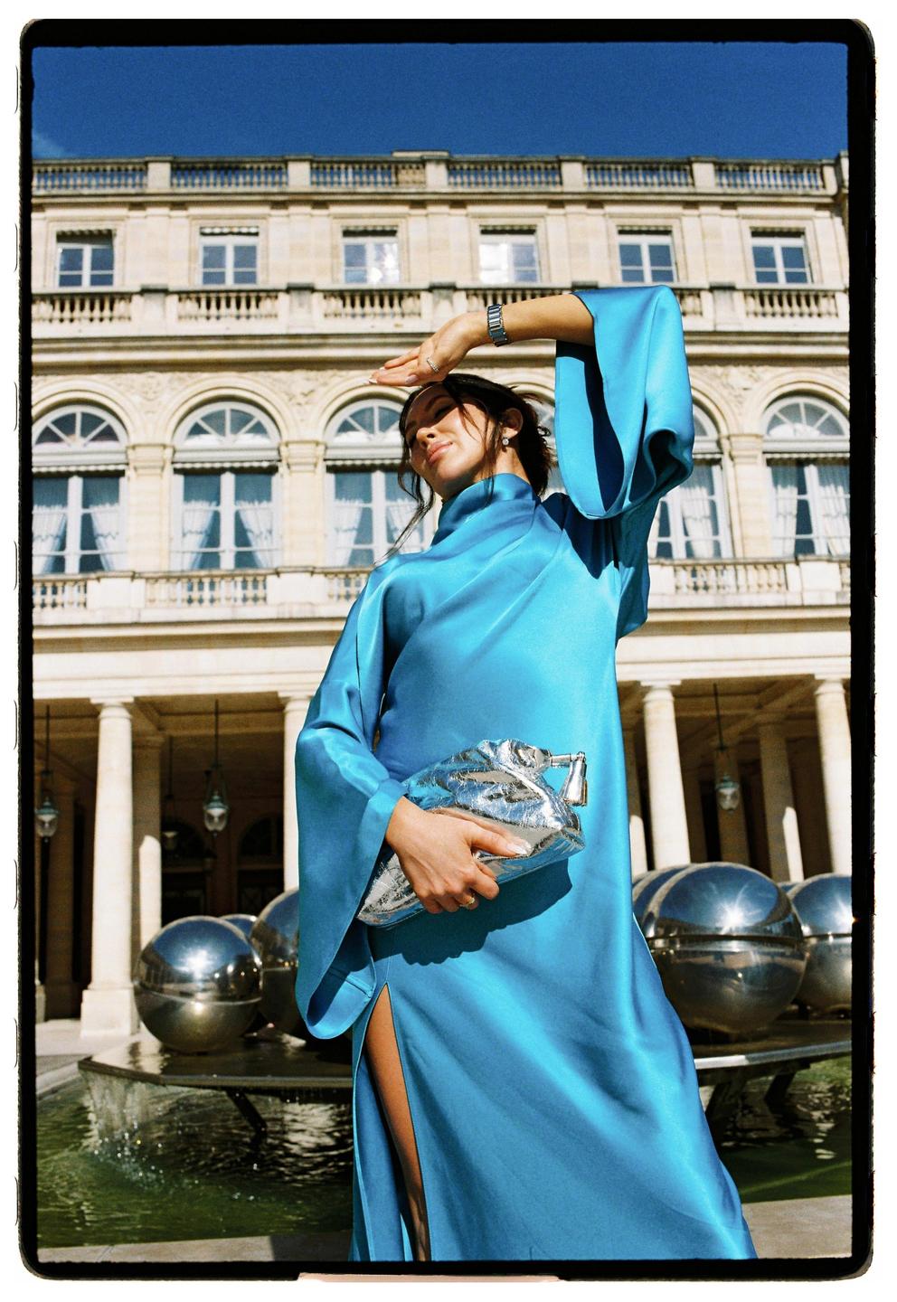 A woman in a blue dress holds a silver clutch while standing in front of a building with a fountain.