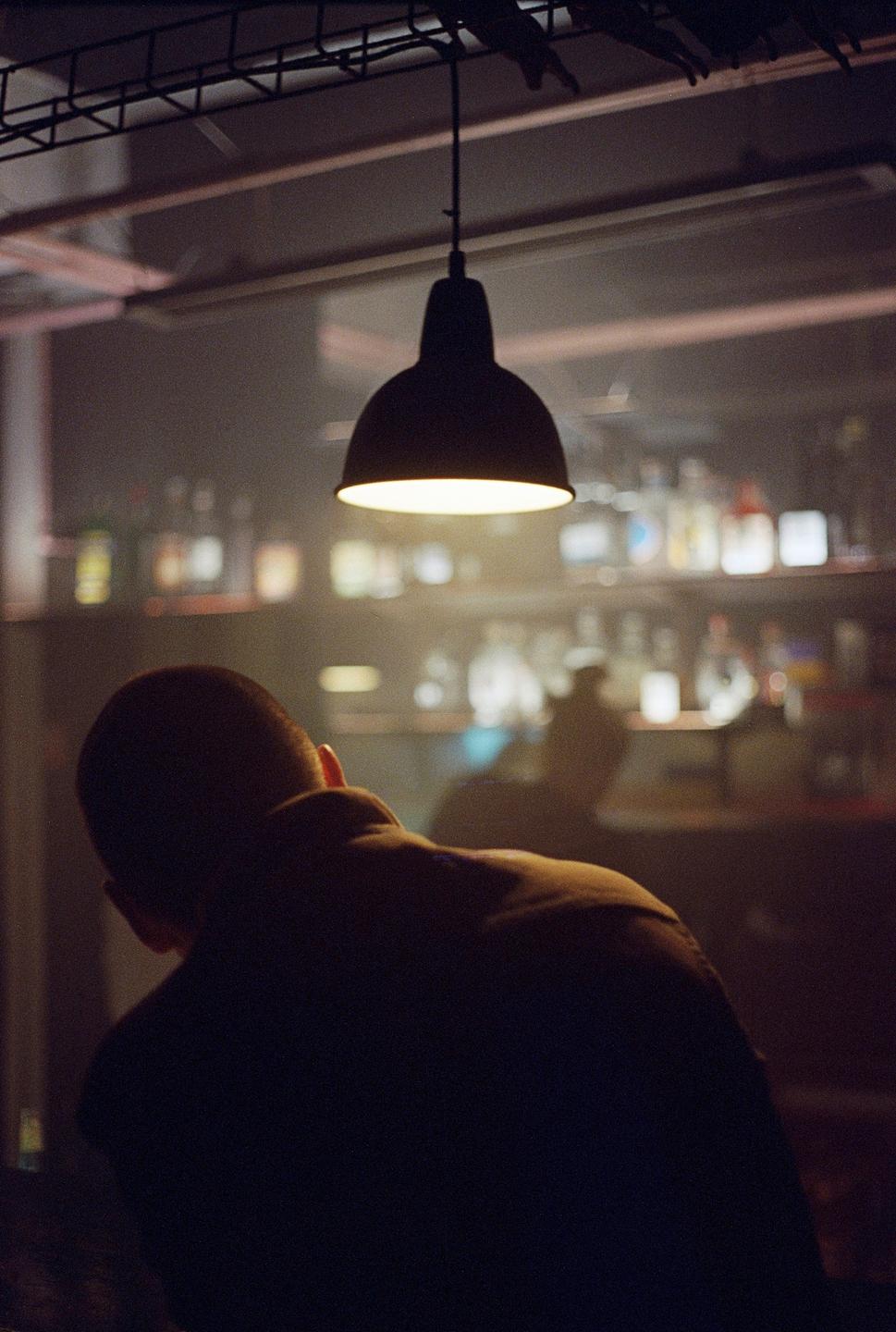 A man sits with his back to the camera under a hanging lamp in a dimly lit bar.