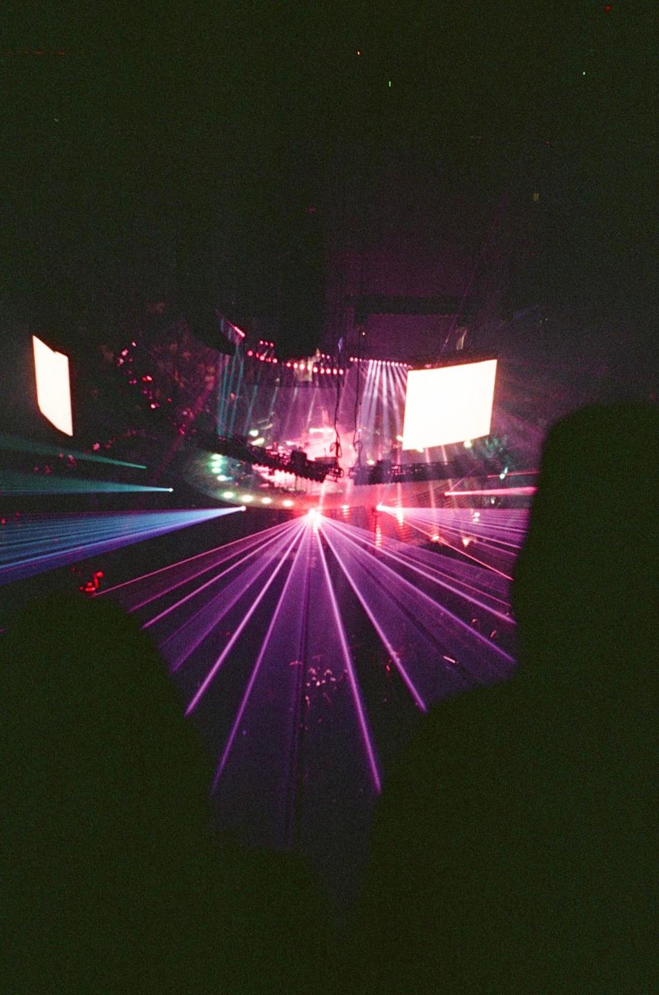 Purple laser beams illuminate a concert stage with a large screen and performers.