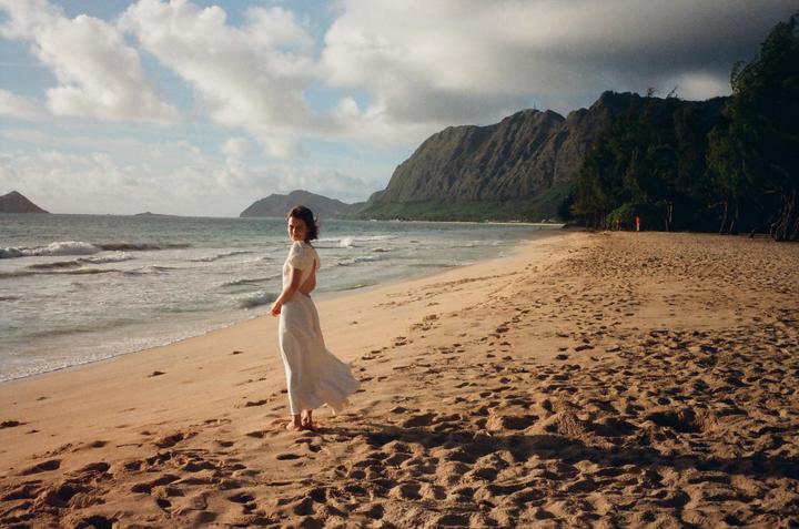 A woman in a white dress stands on a sandy beach with mountains and ocean in the background.