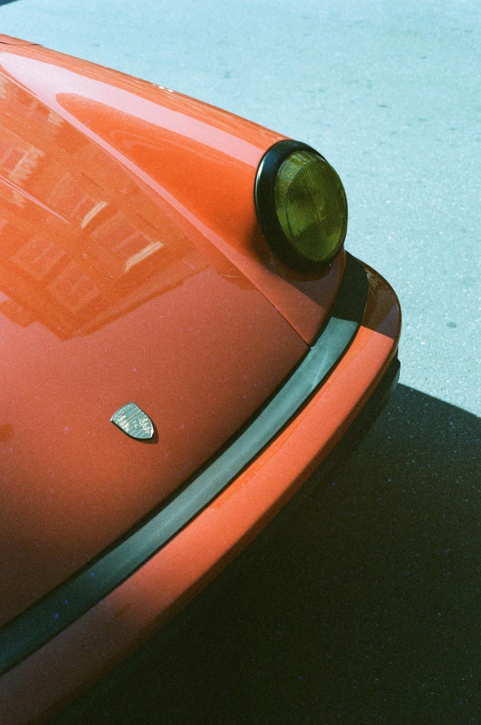 A close-up shot shows the front of an orange classic car with a yellow headlight and a small emblem on the hood.