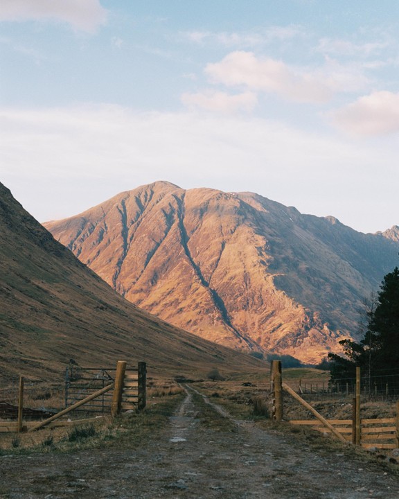 A dirt road leads through a valley towards a large mountain illuminated by golden light.