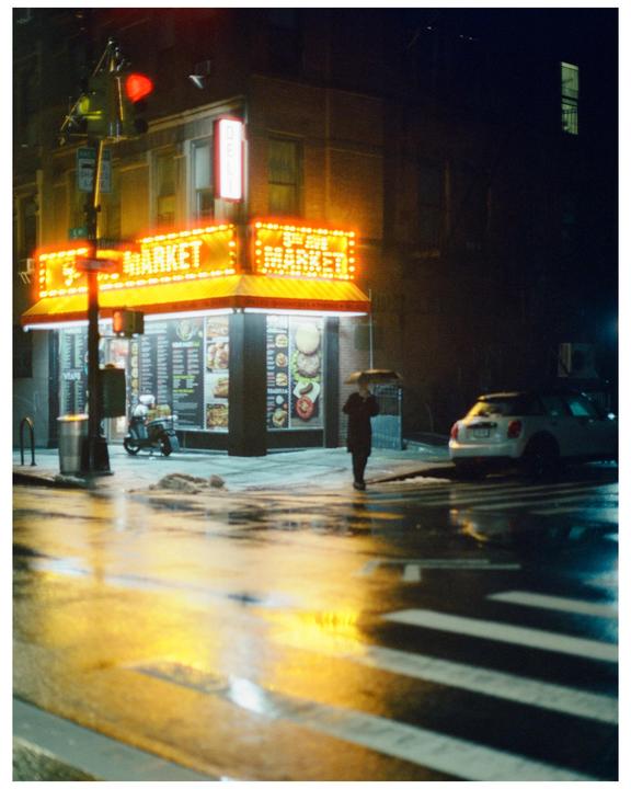 A person with an umbrella walks past a brightly lit market on a wet street at night.