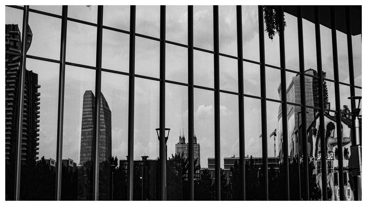 A cityscape with tall buildings and trees is reflected in a grid of windows.