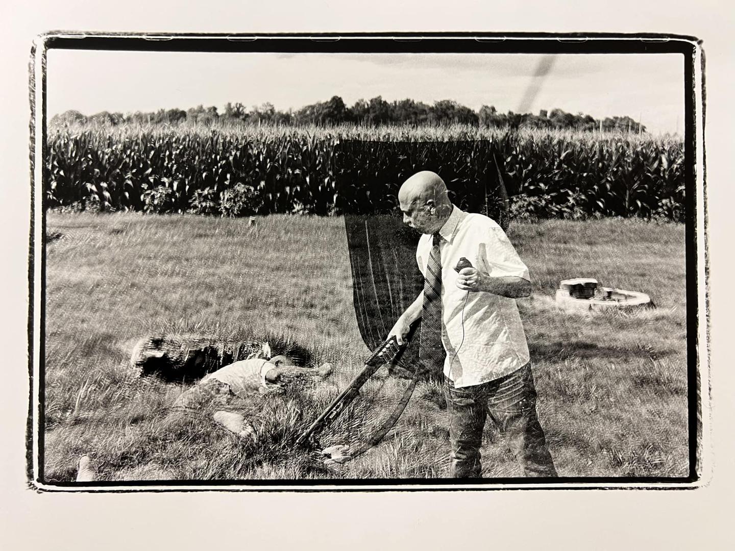 A man in a shirt and tie holds a weapon while standing over a body in a field next to a cornfield.