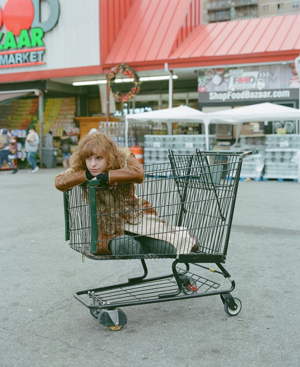 A woman with curly hair sits inside a shopping cart in front of a Food Bazaar Market.