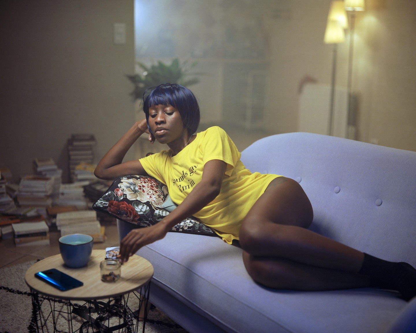 A woman with blue hair and a yellow shirt reclines on a purple sofa next to a small table with a phone and a cup.