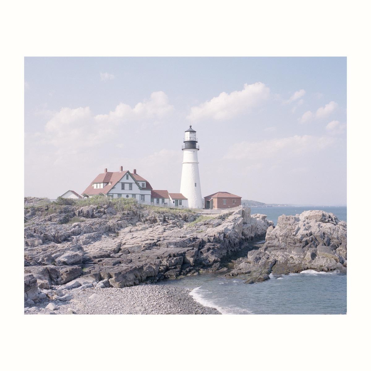 A white lighthouse and buildings with red roofs stand on a rocky coast next to the ocean under a cloudy sky.