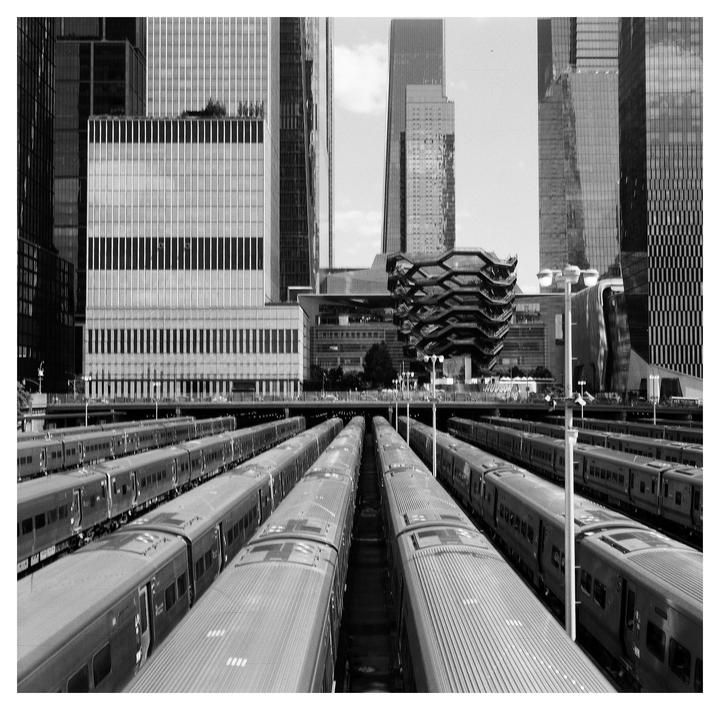 A train yard with multiple tracks and trains is visible in the foreground, with modern skyscrapers and the Vessel structure in the background.