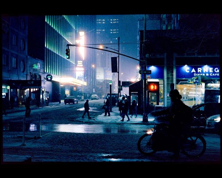 A street scene at night shows people walking and a person on a bicycle, with illuminated buildings and falling snow.