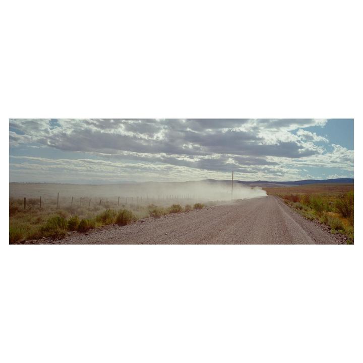 A long dirt road stretches into the distance under a cloudy sky, with a plume of dust rising from a vehicle in the far distance.