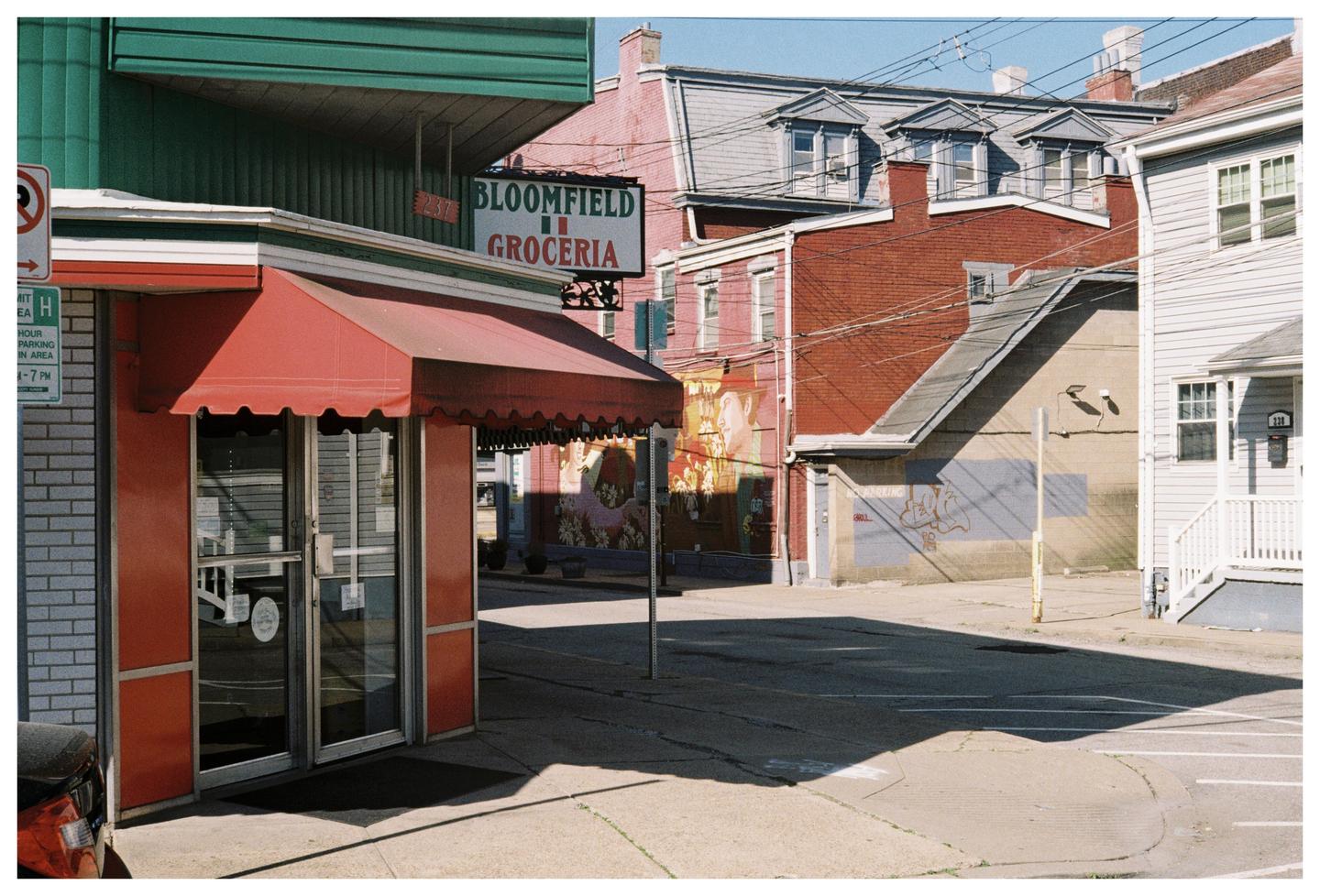A street corner features a grocery store with a red awning and a sign that reads "Bloomfield Groceria," with residential buildings and a mural visible in the background.