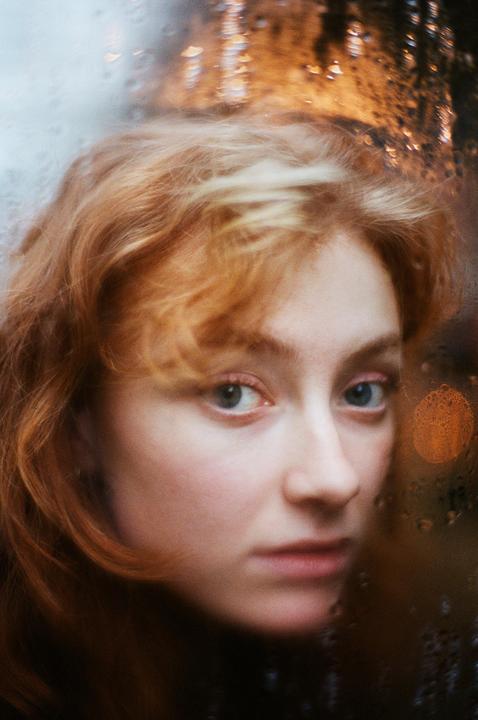A woman with red hair looks through a rain-streaked window at night.