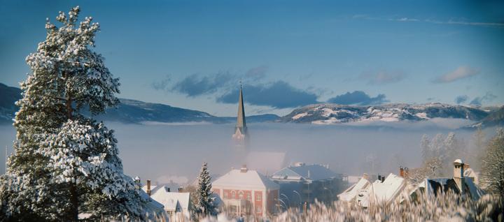 A snow-covered town with a prominent church steeple is nestled in a valley filled with fog, surrounded by snow-capped mountains under a blue sky.