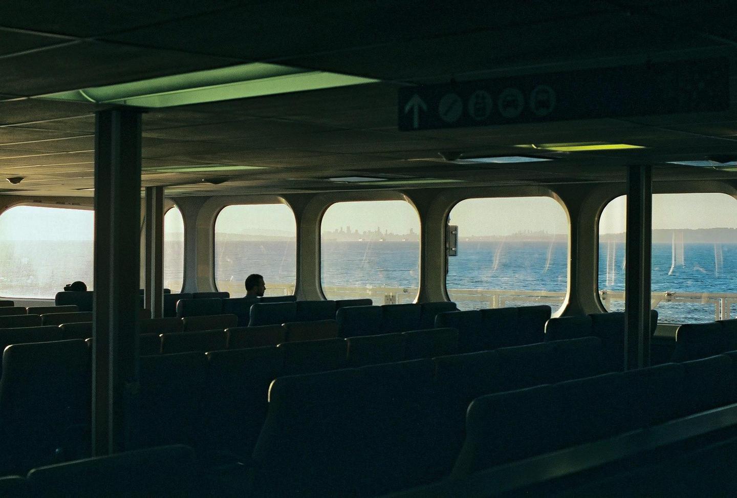 A man sits on a ferry looking out at the water and a distant city skyline.