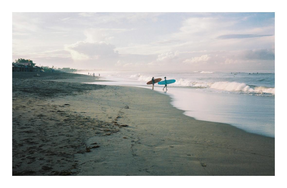 Two surfers walk along a sandy beach with their surfboards as waves crash on the shore.