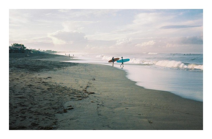Two surfers walk along a sandy beach with their surfboards as waves crash on the shore.